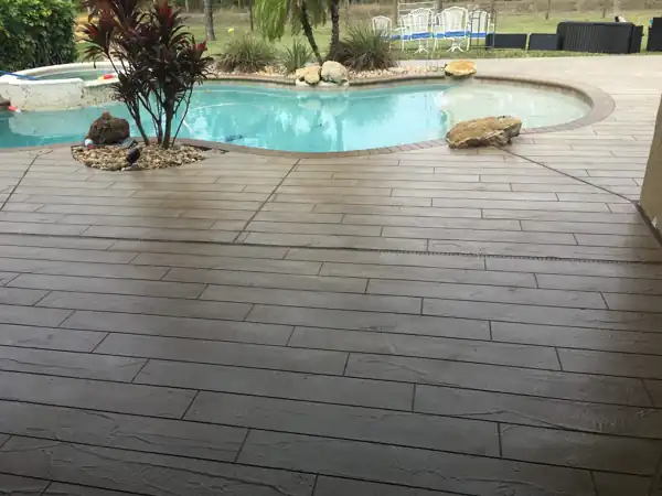 Dark brown wood-plank pattern concrete overlay deck beside a free-form pool with boulder landscaping