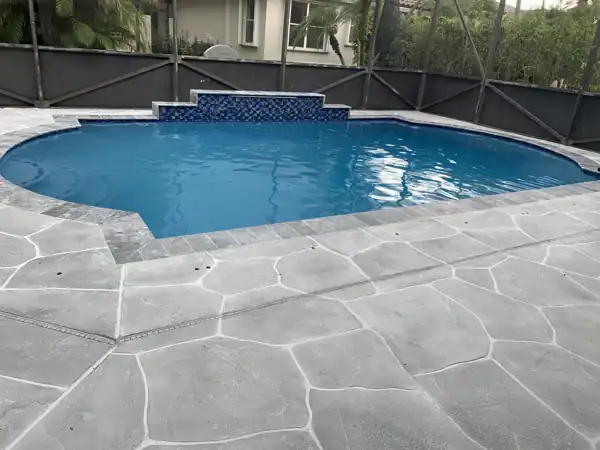 Grey stamped concrete flagstone deck beside a pool with a blue mosaic tile waterfall wall, screened enclosure