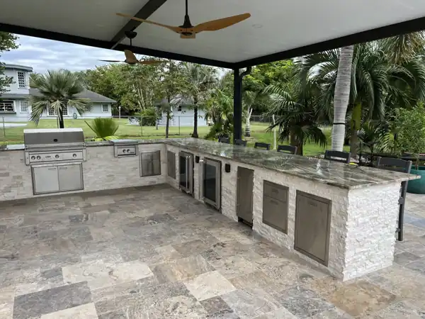 Custom outdoor kitchen with a stacked-stone base and granite countertop under a covered patio, palm trees in the background