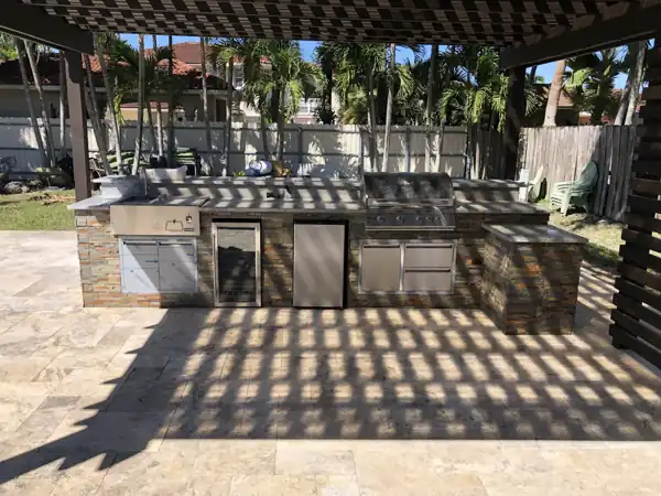 Custom outdoor kitchen with a stone-veneer base and stainless steel grill and appliances under a lattice-covered patio