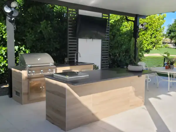Custom outdoor kitchen with light wood-tone cabinetry, black countertop, and stainless steel grill under a covered patio