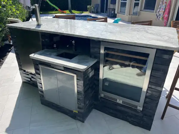 Custom outdoor kitchen bar with dark stone veneer, marble-look countertop, and built-in beverage fridge near the pool