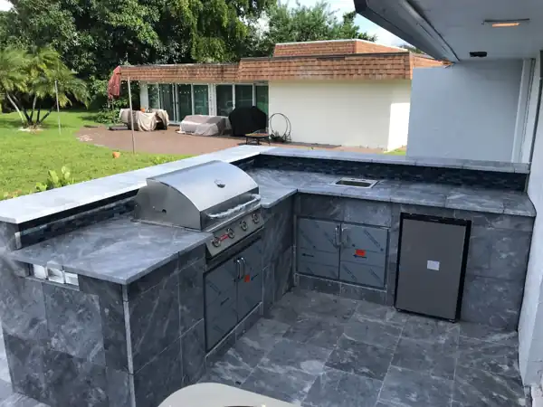 Custom outdoor kitchen with a gray stone-tile counter, built-in grill and refrigerator, under a covered patio