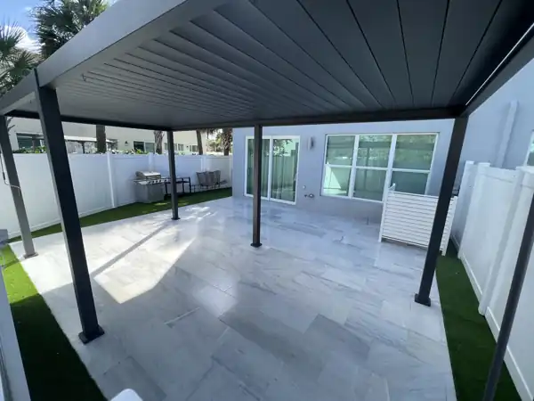 Covered patio with a modern aluminum-paneled roof over a light travertine paver floor, artificial turf border and white vinyl fence