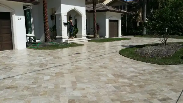 Tan travertine paver driveway leading to a Mediterranean-style home entrance framed by palm trees