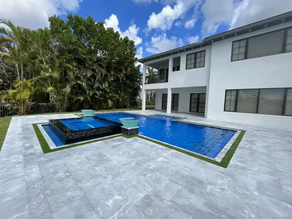 Freeform pool with attached raised spa and turquoise in-pool loungers, light travertine paver deck bordered by turf strips, behind a two-story South Florida home
