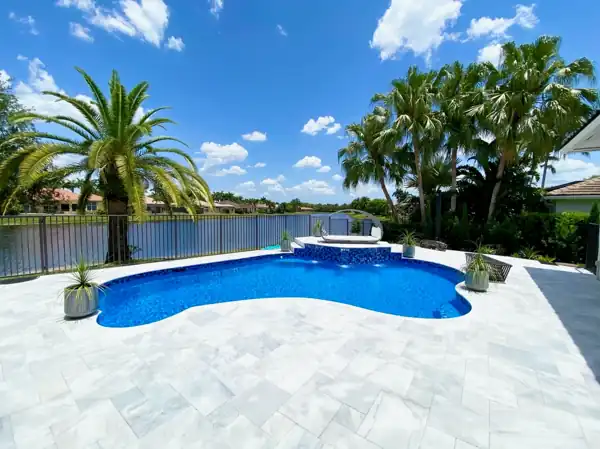 Freeform pool with a raised bubbling spa and travertine paver deck overlooking a South Florida waterway, framed by palm trees