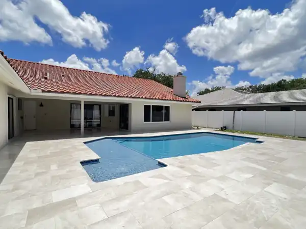 Freeform pool with a tanning ledge and light travertine paver deck behind a terracotta-roof ranch home